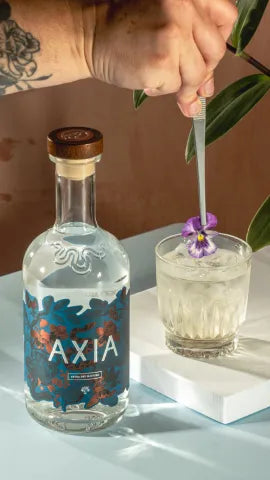 Axia Greek Mastiha Spirit x Gaucho Peach Wine Aristaeus Cocktail on a table with an Axia Spirit bottle next to it and someone adding a flower to the drink
