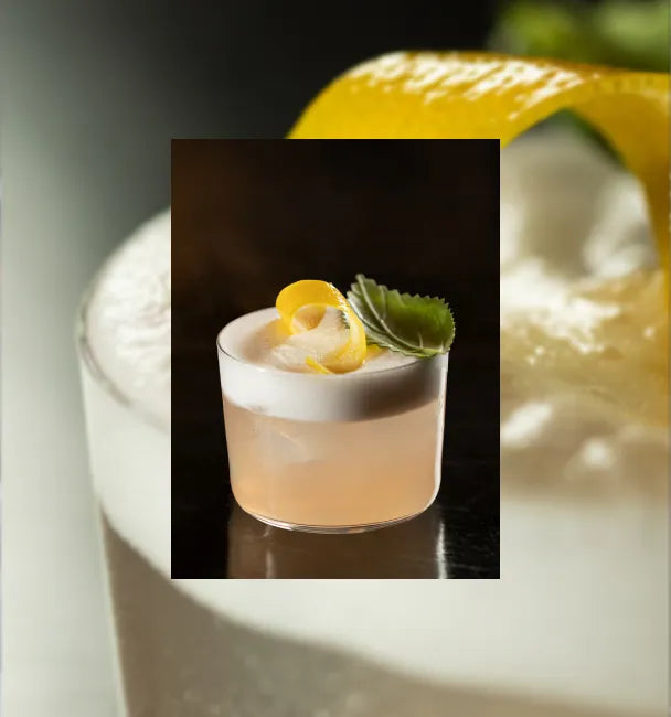 Axia Spirit and Baba au Rum Axia Sour Cocktail on a table with a secondary blurred picture in background of a close-up of another Axia Sour cocktail