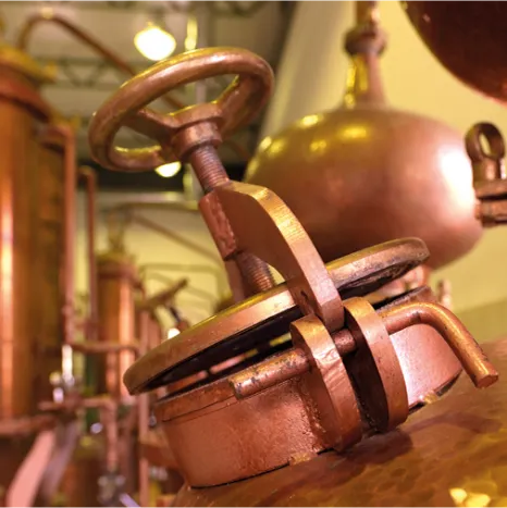 A close-up of a bronze-toned valve with a circular handle and threaded stem, attached to industrial machinery, with blurred copper vessels and pipes in the background for mastiha spirit blending process