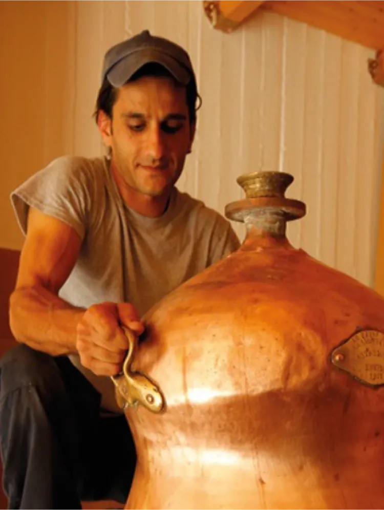A person wearing a gray t-shirt and a cap, crouched down, holding the brass handle of a large copper still for mastiha spirit blending process