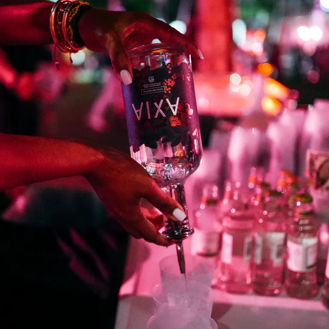 Two hands with white nail polish pouring Axia Mastiha Spirit in a glass on a bar with red light