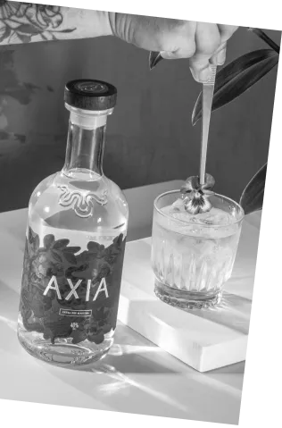 Black and white picture of an Axia Greek Mastiha Spirit x Gaucho Peach Wine Aristaeus Cocktail on a table with an Axia Spirit bottle next to it and someone adding a flower to the drink