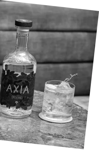 Black and white picture of an Axia Greek Mastiha Spirit and Bar Antoine Four Seasons London Mediterranean Roads Cocktail on a counter next to an Axia Extra Dry Mastiha Spirit bottle