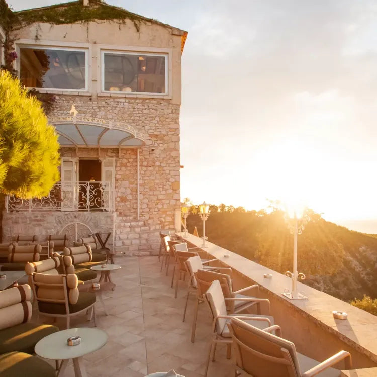 Hôtel Château de la Chèvre D'or, Èze: sunlit terrace with seating overlooks a hillside at sunset, attached to a stone building with large windows
